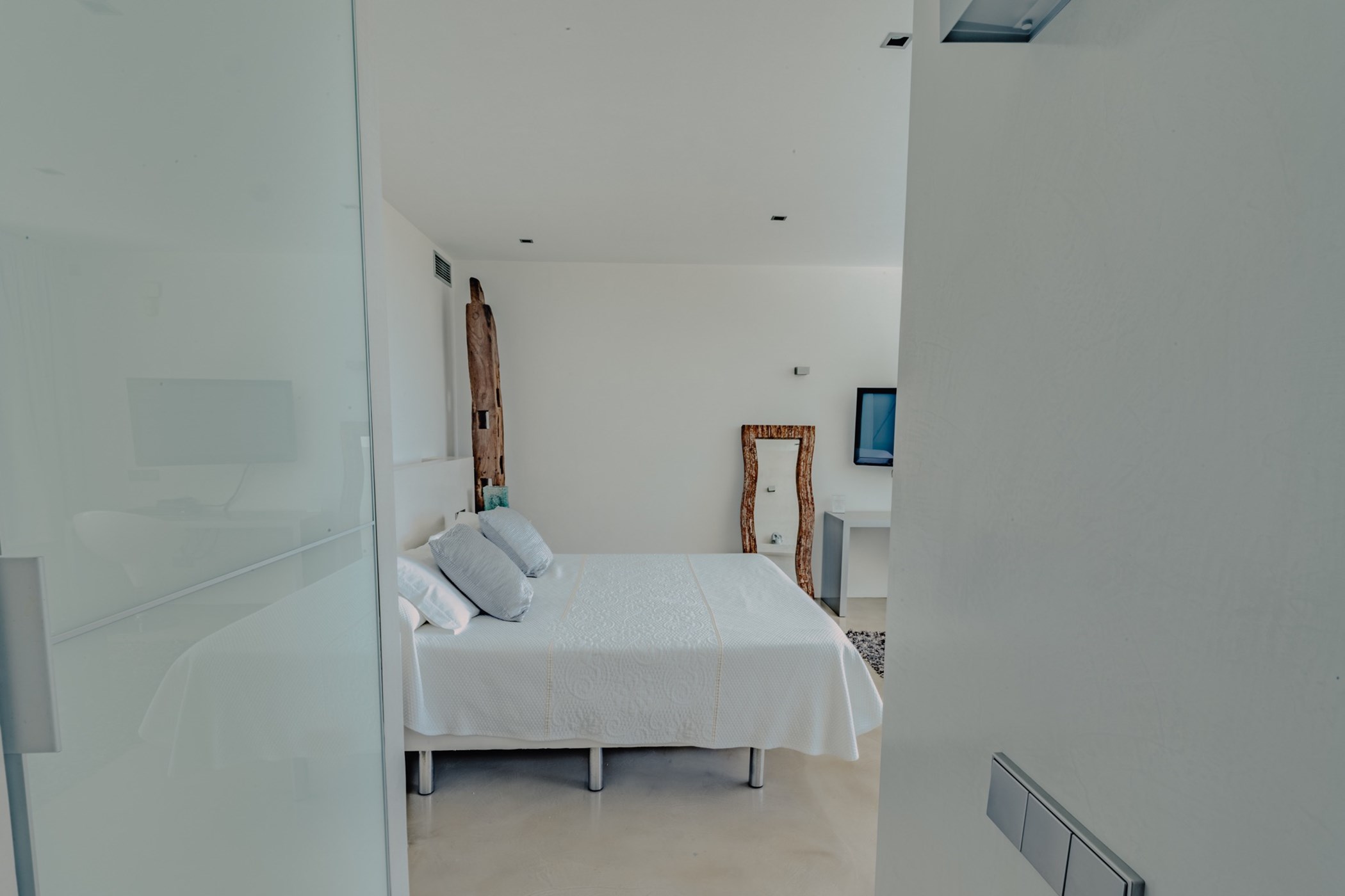 White bedroom interior showing a double bed, wooden mirror and sliding glass door.