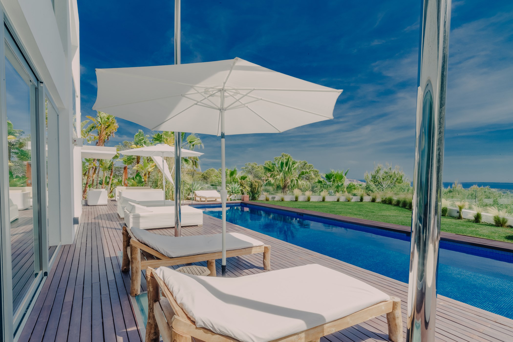 Wooden poolside terrace with white sunbeds, parasols and a flat green lawn area.