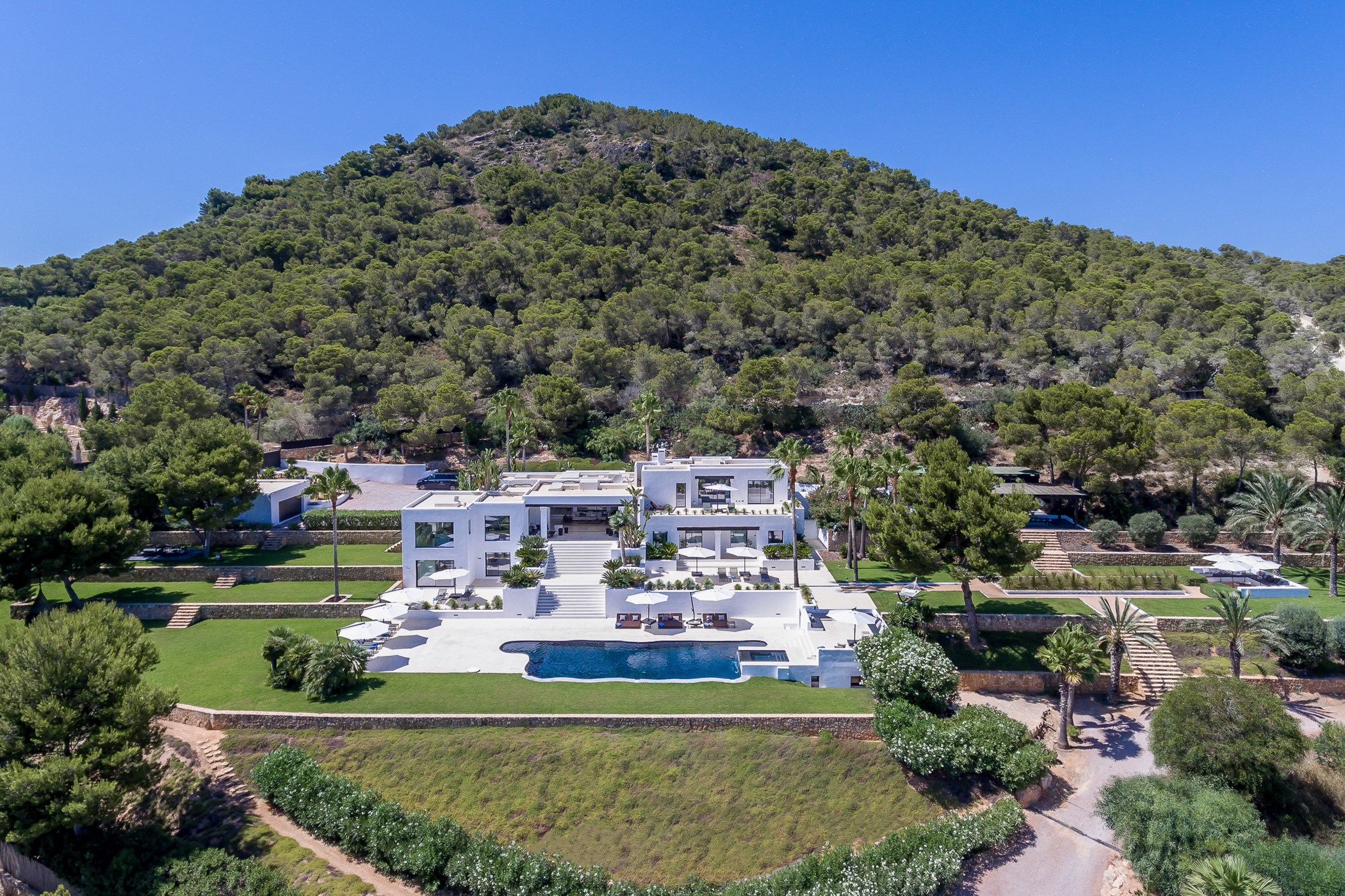 Aerial view of the expansive white modern villa surrounded by green pine-covered hills