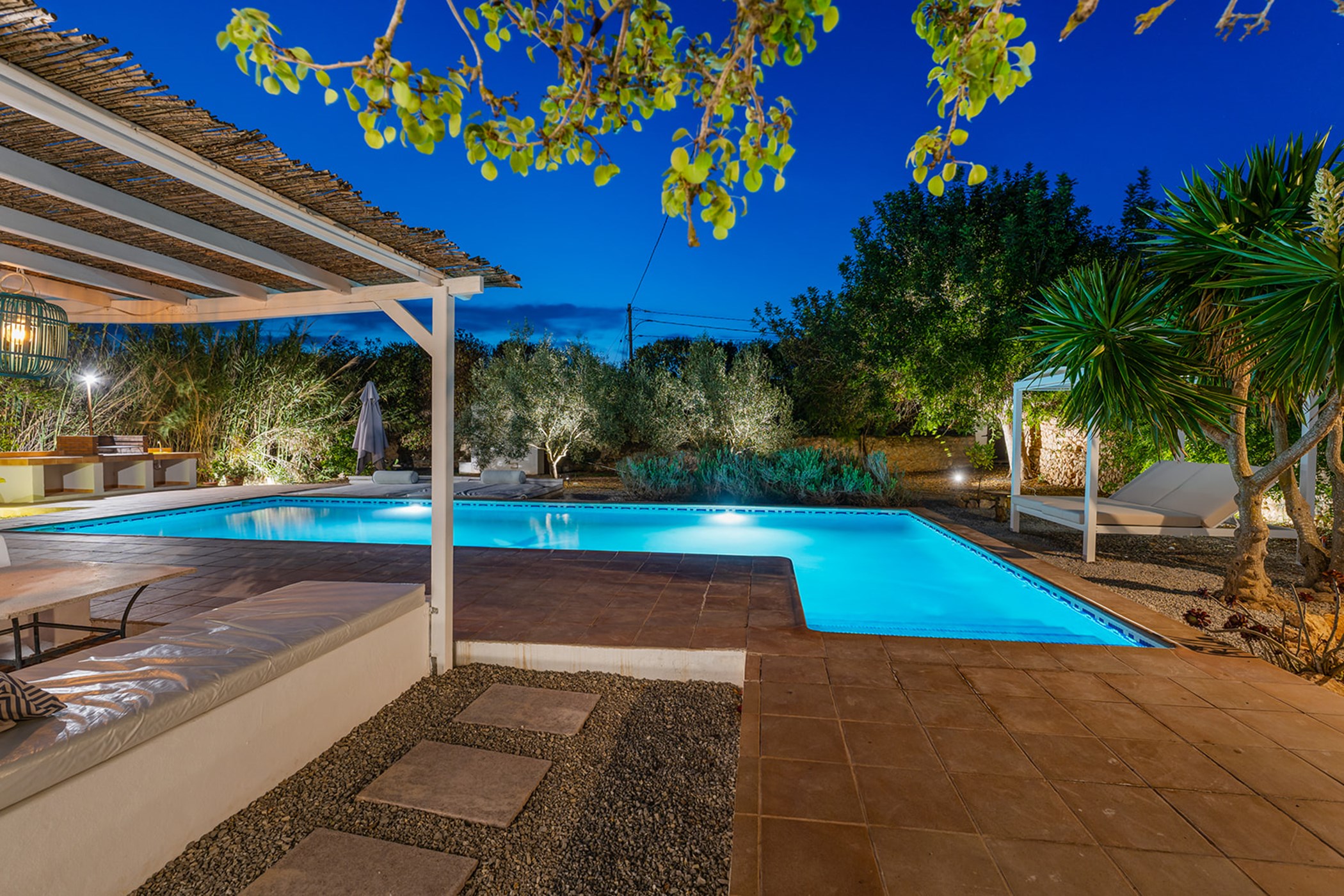 Swimming pool surrounded by trees, a shaded lounge, and a barbecue area at night.