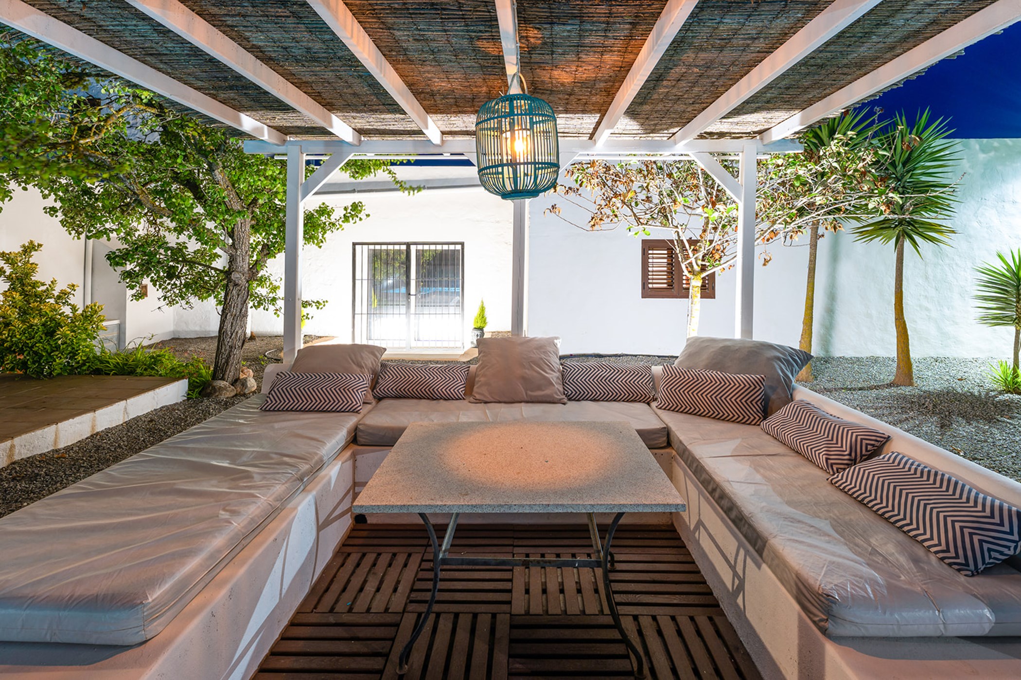 Covered outdoor lounge area with U-shaped seating, striped cushions, and a hanging blue lantern.