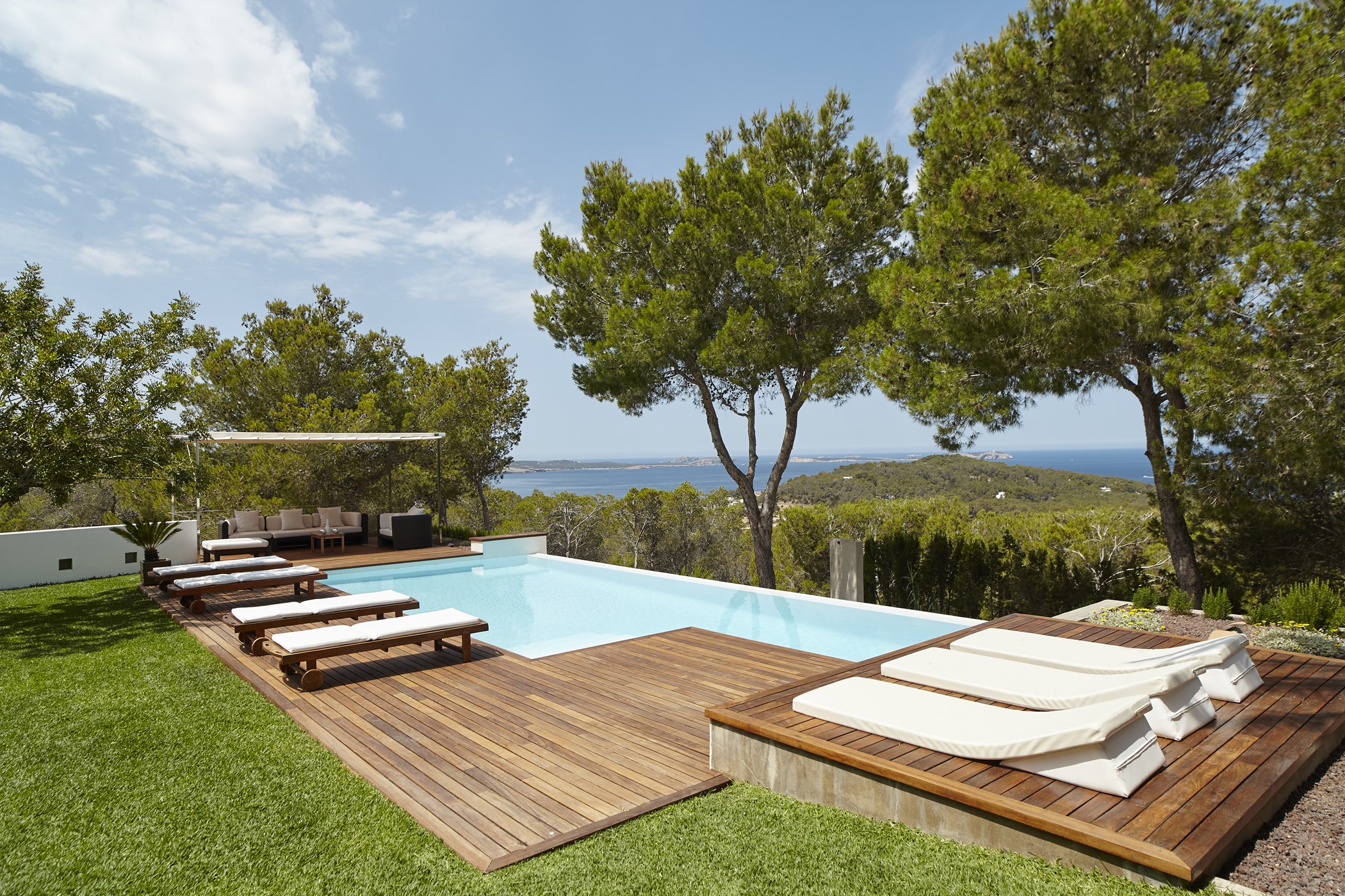 Swimming pool surrounded by wooden decking, sunloungers, green lawn, and mature pine trees.