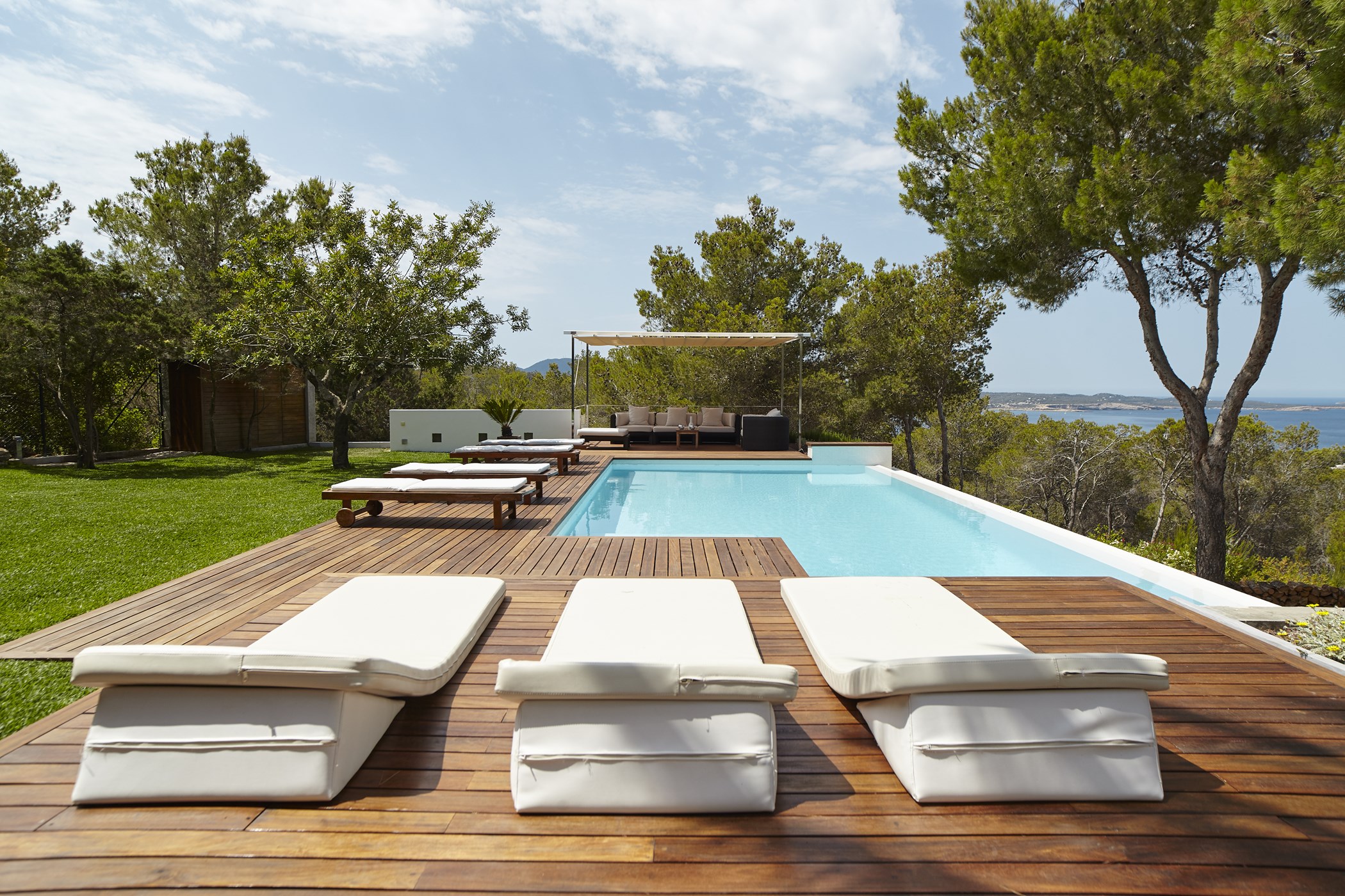 Three white sunloungers on wooden decking beside the swimming pool, facing the sea.